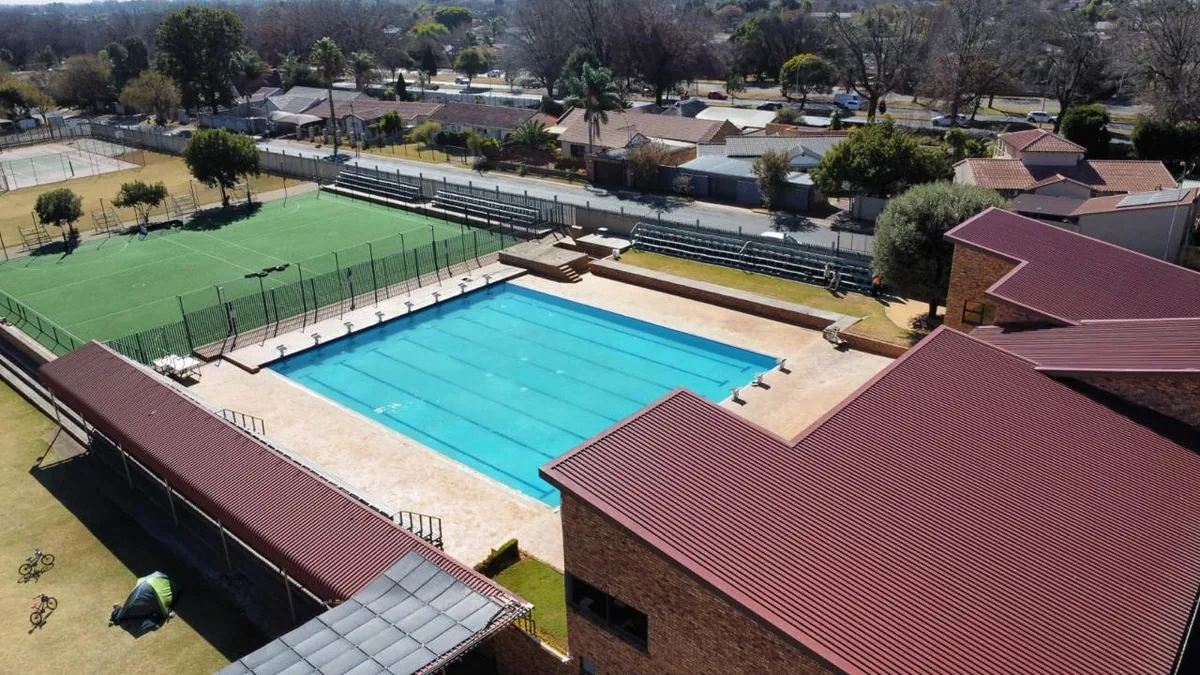 Aerial view of a large institutional lap pool with marked swimming lanes, beside a school sports field and spectator stands